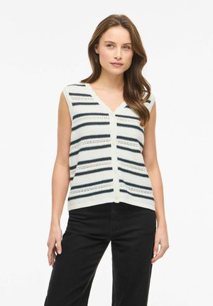 Femme aux cheveux longs et bruns portant un gilet sans manches en maille blanche à rayures horizontales noires et un pantalon noir, debout devant un fond blanc.