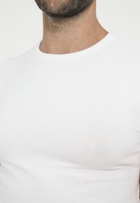 Close-up of a person wearing a plain white crew-neck t-shirt, showing neck and beard details against a white background.
