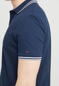 Polo shirt azul marino con un cuello de rayas en contraste, mangas cortas y un logo bordado rojo en el brazo izquierdo. Tejido de algodón suave.