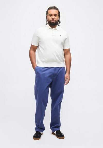 Άντρας με κοτσίδες που φοράει λευκό πόλο με κοντά μανίκια, μπλε παντελόνι και μαύρα αθλητικά παπούτσια, στέκεται με τα χέρια στις τσέπες μπροστά σε μονόχρωμο φόντο.