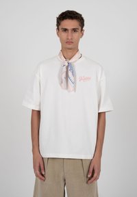 Νεαρός άνδρας που φοράει λευκό φαρδύ T-shirt με την επιγραφή "Απαγορεύεται", μπεζ παντελόνι και ροζ-μπλε ριγωτό μαντήλι στο λαιμό, στέκεται μπροστά σε γκρι φόντο.