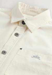 Helles beige Hemd mit Knopfleiste und spitzem Kragen, mit Druckknöpfen und einer Brusttasche mit Etikett. Aus strukturiertem Stoff gefertigt.