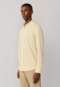 Hombre con camisa de manga larga color amarillo claro abotonada y pantalones beige, de pie frente a un fondo liso gris claro.