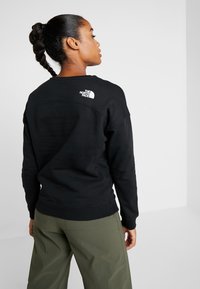 Sweatshirt preto feito de fleece, com decote redondo, punhos canelados e um logotipo branco na parte superior das costas. Combinado com calças verde-azeitona.