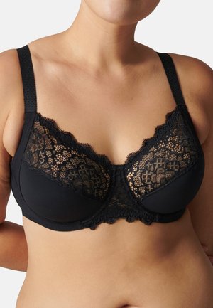 Donna che indossa un reggiseno con ferretto in pizzo nero e mesh con spalline larghe, che mostra il torso superiore e le spalle su uno sfondo neutro.