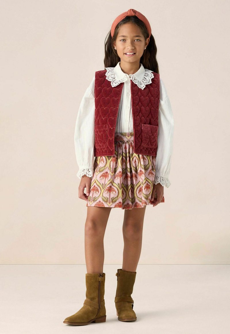 Gilet matelassé rouge avec motif en cœur, blouse blanche avec col en dentelle, jupe rose à motifs et bottines en daim marron. Accent avec un bandeau orange.