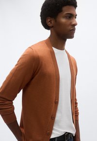 Καφέ ζακέτα με V-λαιμόκοψη, μακριά μανίκια και έξι κουμπιά στο μπροστινό μέρος. Φορεμένη πάνω από λευκό T-shirt, διαθέτει λεία υφή.