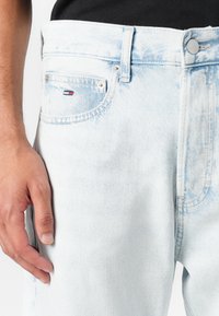 Jean en denim bleu clair avec une finition délavée, design à cinq poches et une petite étiquette logo sur la poche avant. Fermeture à bouton à la taille.