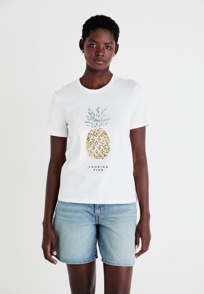 T-shirt bianca in cotone con una grafica di ananas dorato dai dettagli verdi e il testo "LOOKING PINE" sottostante, abbinata a pantaloni shorts in denim chiaro.