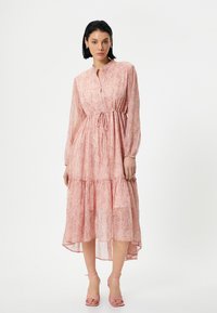 Roze tiered jurk met lange mouwen, knoopsluiting aan de voorkant, elastische taille en delicate bloemenprint. Gekozen met roze bandjeshakken.