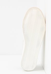 Semelle d'une basket blanche pressée contre un mur blanc, montrant un motif de bande en chevrons.