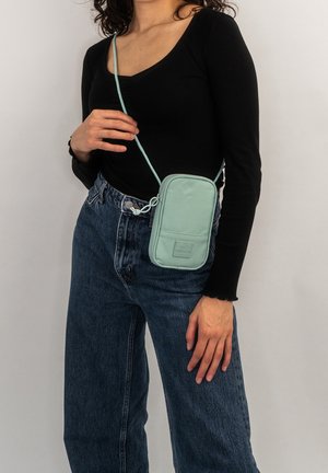 Donna che indossa un top nero a maniche lunghe e jeans blu, mentre porta una piccola borsa a tracolla verde chiaro con una tasca frontale.