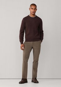 Mann, der einen dunkelbraunen Pullover, beige Hosen und dunkelbraune Lederschuhe trägt, steht vor einem einfarbigen, hellen Hintergrund.