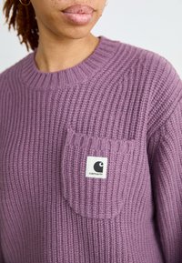 Suéter de punto morado con textura acanalada, que presenta un pequeño bolsillo en el pecho y un parche con el logo de Carhartt. Cuello redondo.