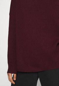 Maglione a maglia borgogna con una vestibilità ampia, maniche lunghe e polsini a coste, abbinato a pantaloni neri. Tessuto morbido con un design semplice.