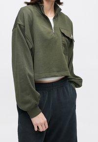 Personne portant un sweat court à quart de zip vert olive avec une poche poitrine à rabat, un pantalon de survêtement noir et un t-shirt blanc en dessous, debout de manière décontractée.