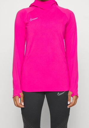 Femme portant un haut de sport rose vif à manches longues avec des trous pour les pouces et un legging noir avec des logos Nike sur les deux vêtements.