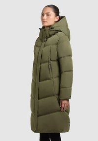 Olive roheline pikk puffjakker, millel on kapuuts, tikkitud osad, eesne lukuga ja küljetaskud. Sile tekstuur ja keskmine kaal.