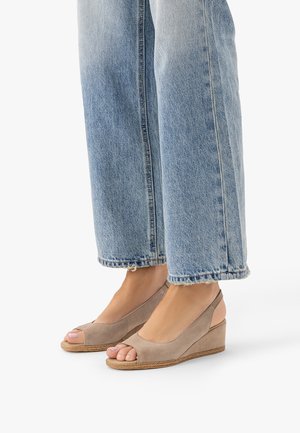 Stopy w beżowych zamszowych sandałach na klinie z odkrytymi palcami, zestawione z jasnoniebieskimi dżinsami z postrzępionym brzegiem na gładkim tle.