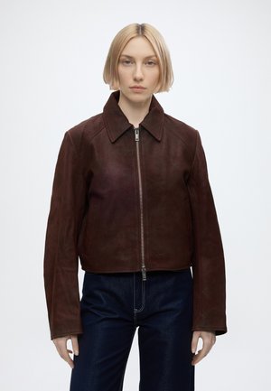 Mujer con cabello rubio corto que lleva una chaqueta de ante marrón oscuro con cremallera y pantalones azul oscuro, de pie sobre un fondo claro y liso.