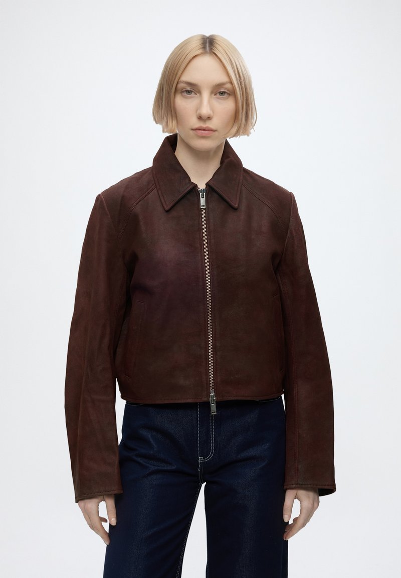 Femme aux cheveux blonds courts portant une veste zippée en suède marron foncé et un pantalon bleu foncé, debout devant un fond clair uni.