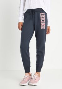 Persoon in marineblauwe joggingbroek met roze "ABERCROMBIE" tekst op het rechterbeen en roze en witte sneakers, staand tegen een effen achtergrond.