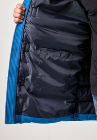 Giacca trapuntata blu con interno in raso blu scuro e tasca interna, mostrante un rivestimento esterno strutturato e cordino in fondo.