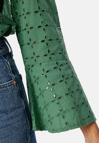 Blouse verte à manches évasées, ornée de motifs découpés brodés et d'une finition texturée. Associée à un jean pour un contraste.