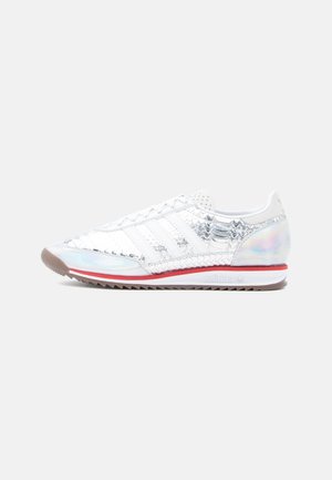 Witte sneaker met iriserende accenten, textuurrijke schubben op het bovenwerk, rood randje aan de zool en klassiek ontwerp met drie strepen. Rubberen buitenzool.