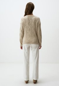 Beige gestrickter Pullover mit einem strukturierten, offenen Muster, Rundhalsausschnitt und langen Ärmeln, kombiniert mit weißen Hosen und braunen Schuhen.