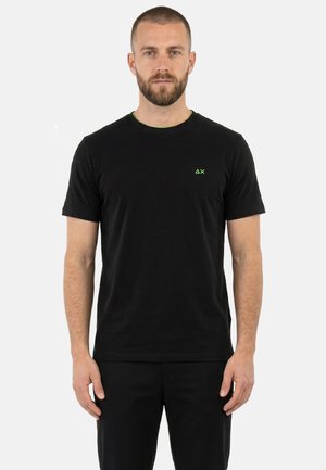 Uomo barbuto con capelli corti che indossa una maglietta nera a maniche corte con un piccolo logo verde ΔX sul petto a sinistra, in piedi dritto.