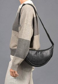 Bolso de hombro de cuero negro con forma curva, que presenta una textura suave y un diseño minimalista, llevado sobre un suéter de rayas beige y gris.