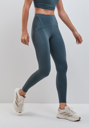 Donna con leggings blu a vita alta e scarpe da corsa bianche che cammina su uno sfondo chiaro uniforme.