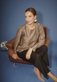Donna con capelli castani, vestita con una camicetta metallica, pantaloni neri e tacchi luccicanti, seduta su una sedia in pelle marrone contro uno sfondo blu.