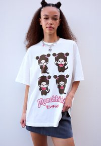 Mlada ženska z kodrastimi lasmi in dvema bunama nosi belo oversize majico z motivom štirih risanih Monchhichi junakov in kratko sivo krilo.