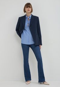 Woman standing against white wall wearing navy blazer, blue striped shirt, dark blue flare jeans, and beige pointed heels.