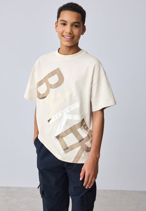 Lächelnder Junge trägt ein beiges oversized T-Shirt mit großem "BAKER"-Aufdruck und schwarze Cargohosen, steht vor einem schlichten hellen Hintergrund.