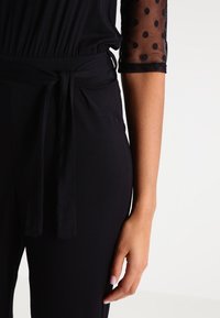 Zwarte jumpsuit met een aansluitende taille, een gestrikt stoffen riempje en doorschijnende zwarte stippenmouwen, met een gladde afwerking en een ontspannen pasvorm.