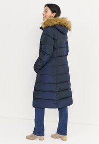 Marineblauwe pufferjas met afneembare nepbontkraag, verticale naden en zijzakken. Draagt over lichtblauwe spijkerbroeken en beige enkellaarzen.