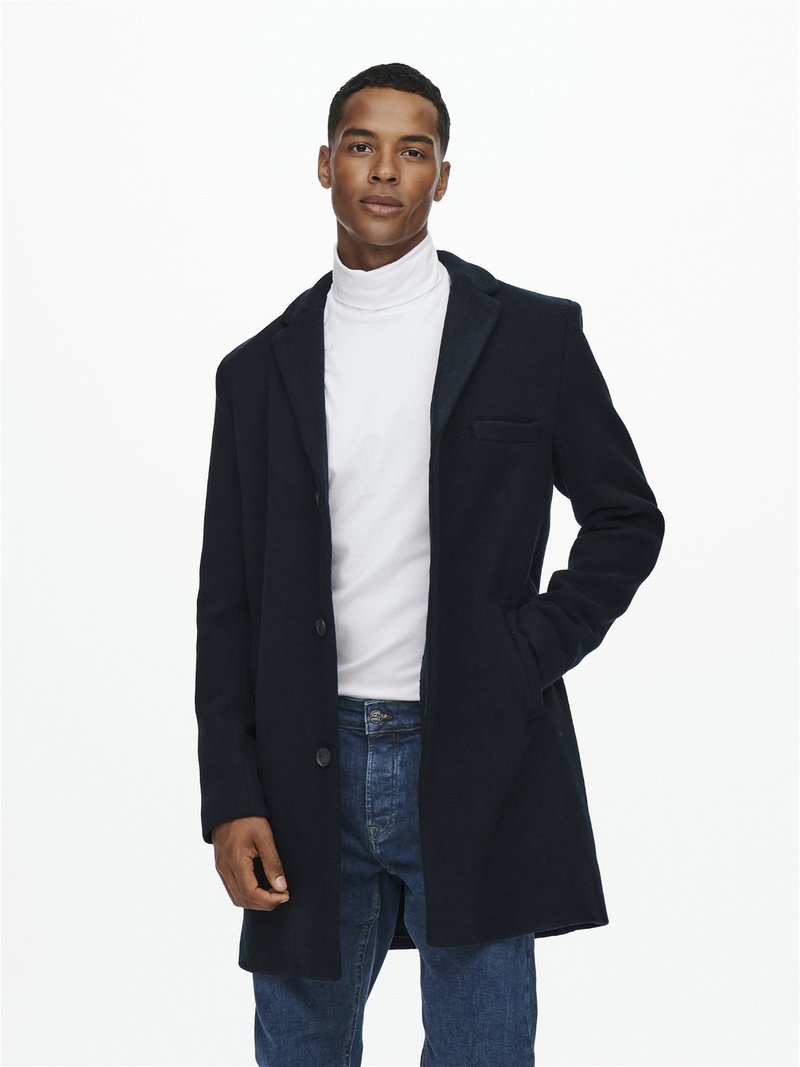 Manteau en laine noir avec un col cranté, trois boutons et des poches latérales, porté sur un pull à col roulé blanc. Un jean bleu simple complète la tenue.