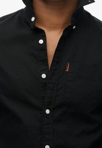 Camicia nera con bottoni in cotone, con colletto, dotata di bottoni bianchi e una piccola etichetta rossa sulla tasca. Superficie strutturata.