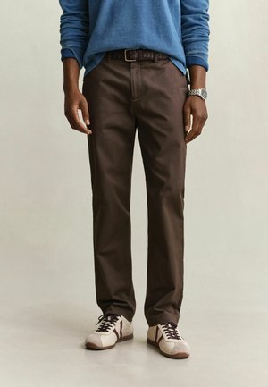 Homme portant un pantalon marron, des baskets crème et marron, une chemise bleue à manches longues, une ceinture tressée marron et une montre-bracelet argentée, debout sur un sol clair.