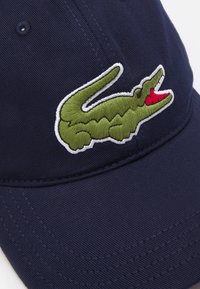 Jūras zila cepure ar izšūtu zaļu krokodila logotipu ar sarkaniem akcentiem priekšā. Izgatavota no izturīga, teksturēta auduma.