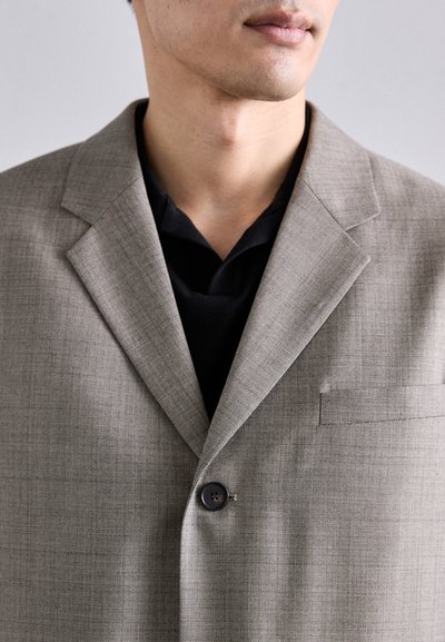 Hellgrauer taillierter Blazer aus strukturiertem Stoff, mit einem Ein-Knopf-Verschluss und einer Brusttasche, getragen über einem schwarzen Hemd.