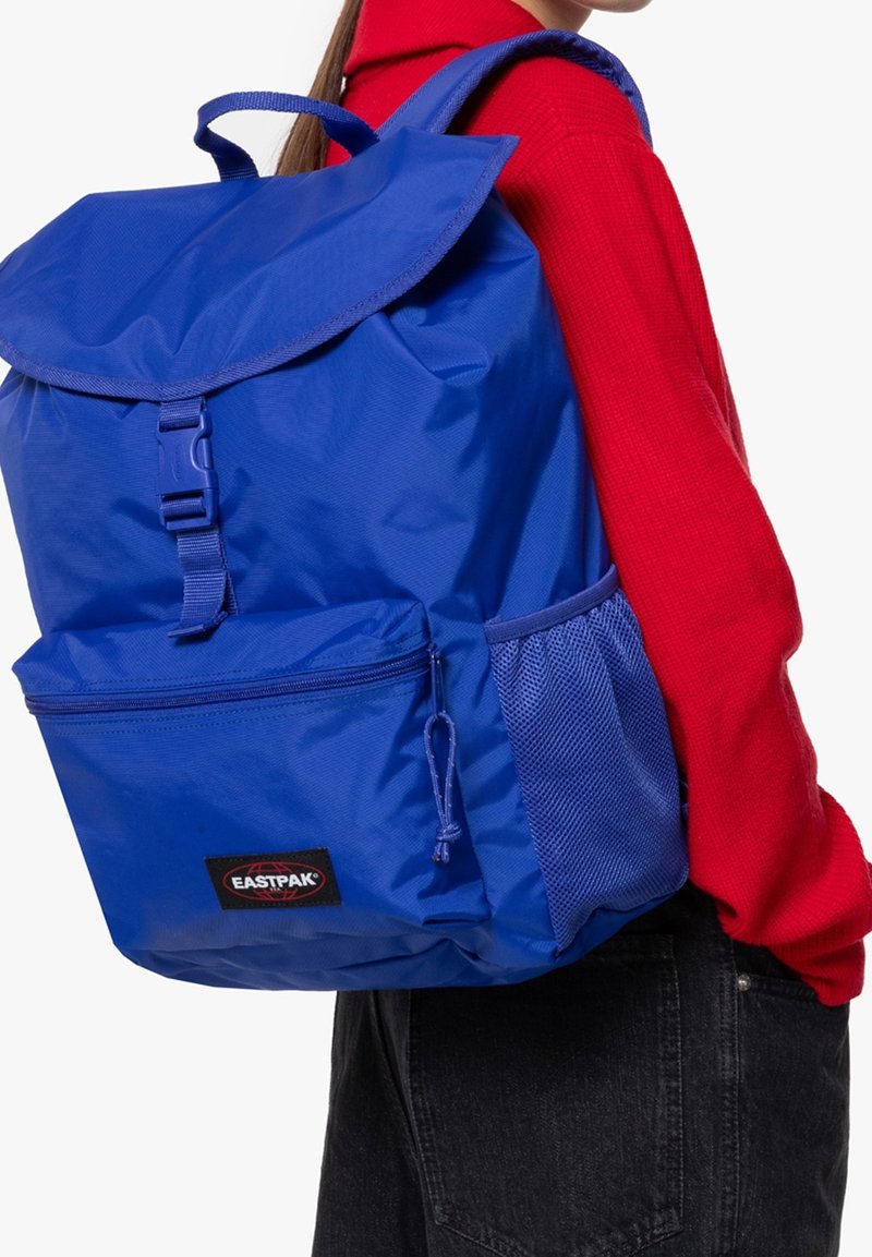 Personne portant un sac à dos Eastpak bleu par-dessus un pull rouge et un jean noir, vue de côté et de dos sur fond blanc.