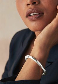 Bracciale in metallo argentato, design curvo con una texture lucida, indossato su un polso su uno sfondo neutro.