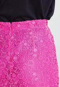 Detailní záběr na živě růžovou krajkovou sukni s květinovým vzorem a zipem, která je nošena s černým topem.