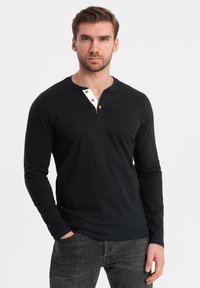 Mies, jolla on lyhyt ruskea hius ja parta, pukeutuneena mustaan pitkähiharaiseen henley-paitaan, jossa on puunapit, ja tummanharmaisiin farkkuihin, seisoo yksi käsi taskussa.