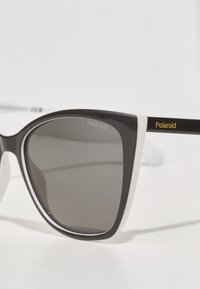 Occhiali da sole cat-eye bianco e nero con lenti grigie. Presentano una finitura lucida, il marchio dorato "Polaroid" sull'asta e una texture uniforme.