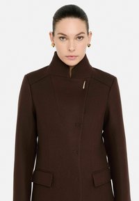 Cappotto in lana bordeaux con colletto alto, design minimalista, spilla dorata singola e due tasche frontali. Texture liscia.